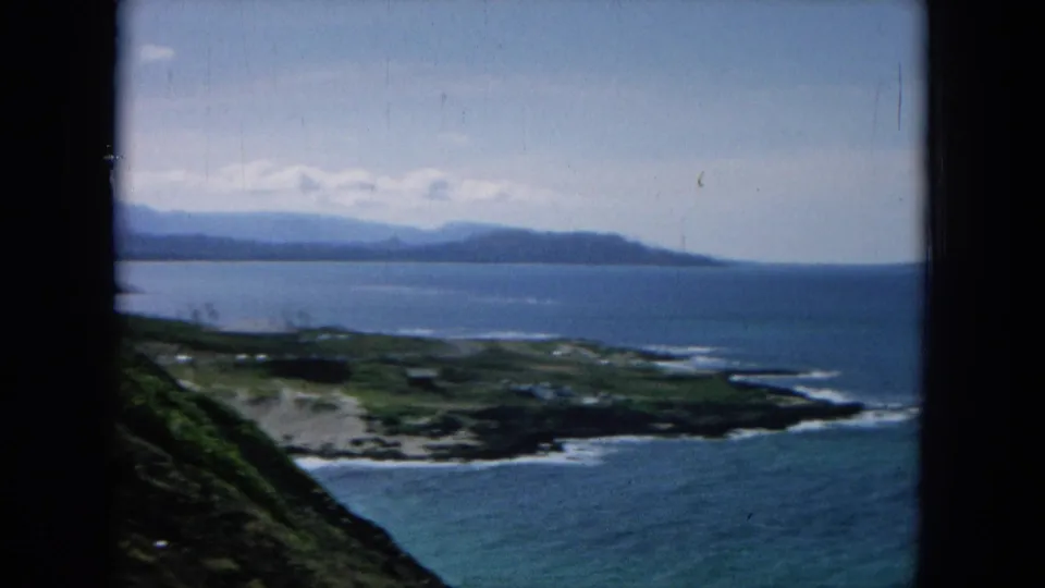 1964 8mm Hawaiian coastal landscape with a blue bay, green hillside foreground, surf on the shore, and distant mountain ridges.
