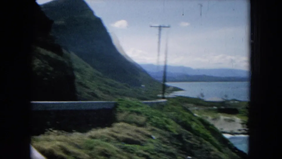 1964 8mm Hawaiian roadside view with a steep green cliff, utility pole, ocean below, and distant ridges across the water.