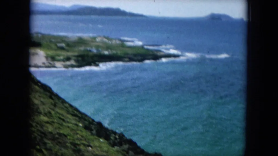 1964 Hawaii home-movie frame looking over blue ocean, surf, distant shore, and a green coastal slope.