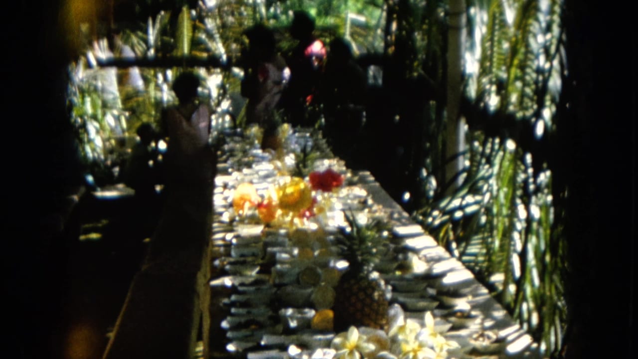 1964 Hawaii home-movie frame of a long garden banquet table with place settings, fruit, flowers, and shadowed people nearby.