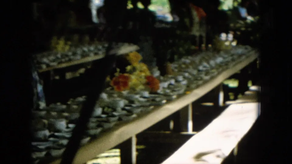 1964 Hawaii home-movie frame showing a shaded outdoor banquet table with glasses, plates, flowers, fruit, and indistinct people in the background.