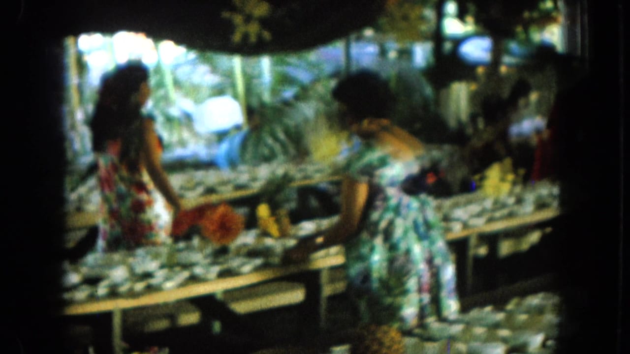 Two women lean over tables lined with small bowls in a shaded outdoor display.