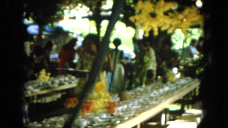 1964 Hawaii home-movie frame with visitors and women around tables of small bowls under tropical tree cover.