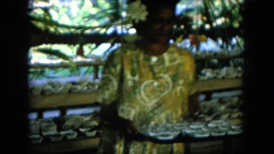 1964 Hawaii frame showing a woman near a table of small bowls, with foliage and fair structures behind her.