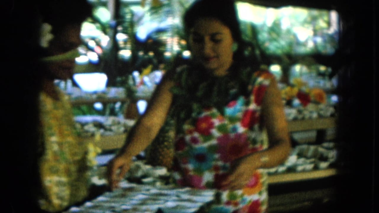 1964 Hawaii home-movie frame showing a woman in a colorful floral dress near a shaded buffet table set with white dishes.