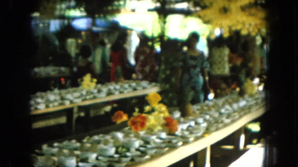 1964 Hawaii home-movie frame showing guests near a long buffet table with white dishes and bright tropical flowers.