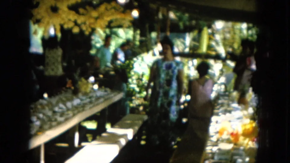 1964 Hawaii home-movie frame with women in patterned dresses beside a long buffet table in a shaded tropical setting.