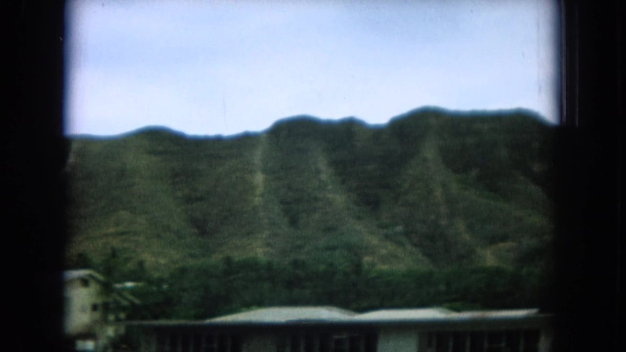 Green mountain ridge seen through a dark window frame above low residential roofs.