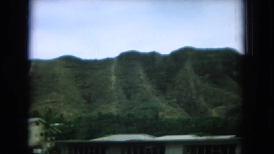 1964 8mm Hawaii home-movie view of a green mountain ridge and low buildings framed by a dark window.