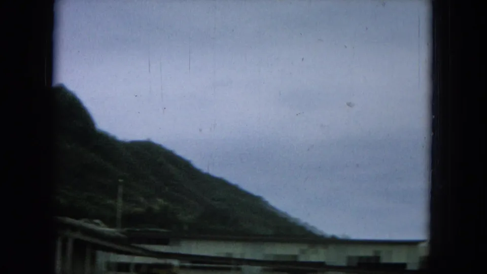 Lower window-pane view of a Hawaiian mountain slope with pale sky and residential rooflines on 8mm film.