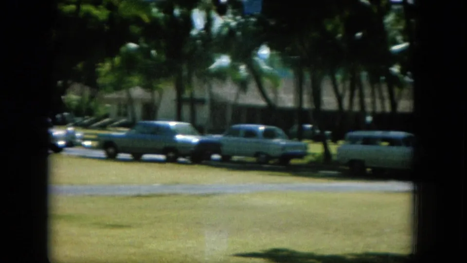 1964 Oahu home-movie view of classic cars parked near palms, lawn, roadway, and a low building.
