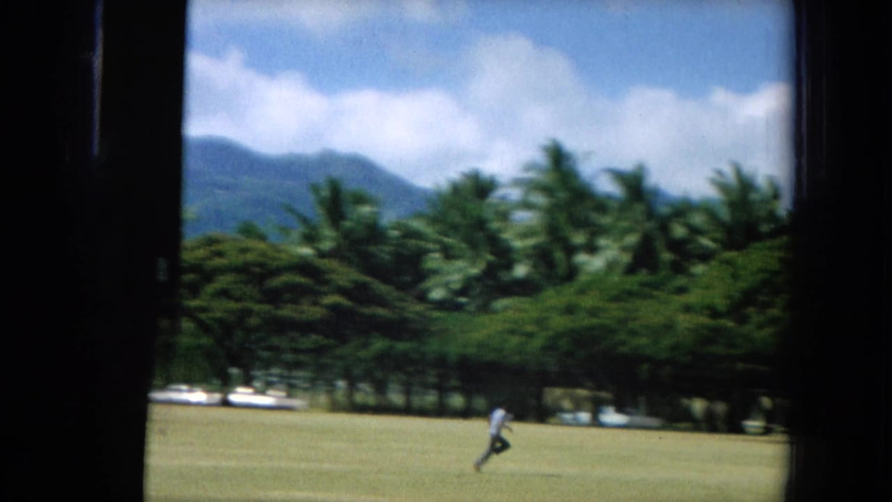 1964 Oahi, Hawaii 8mm frame showing a distant person running across a sunlit field bordered by palms and mountains.