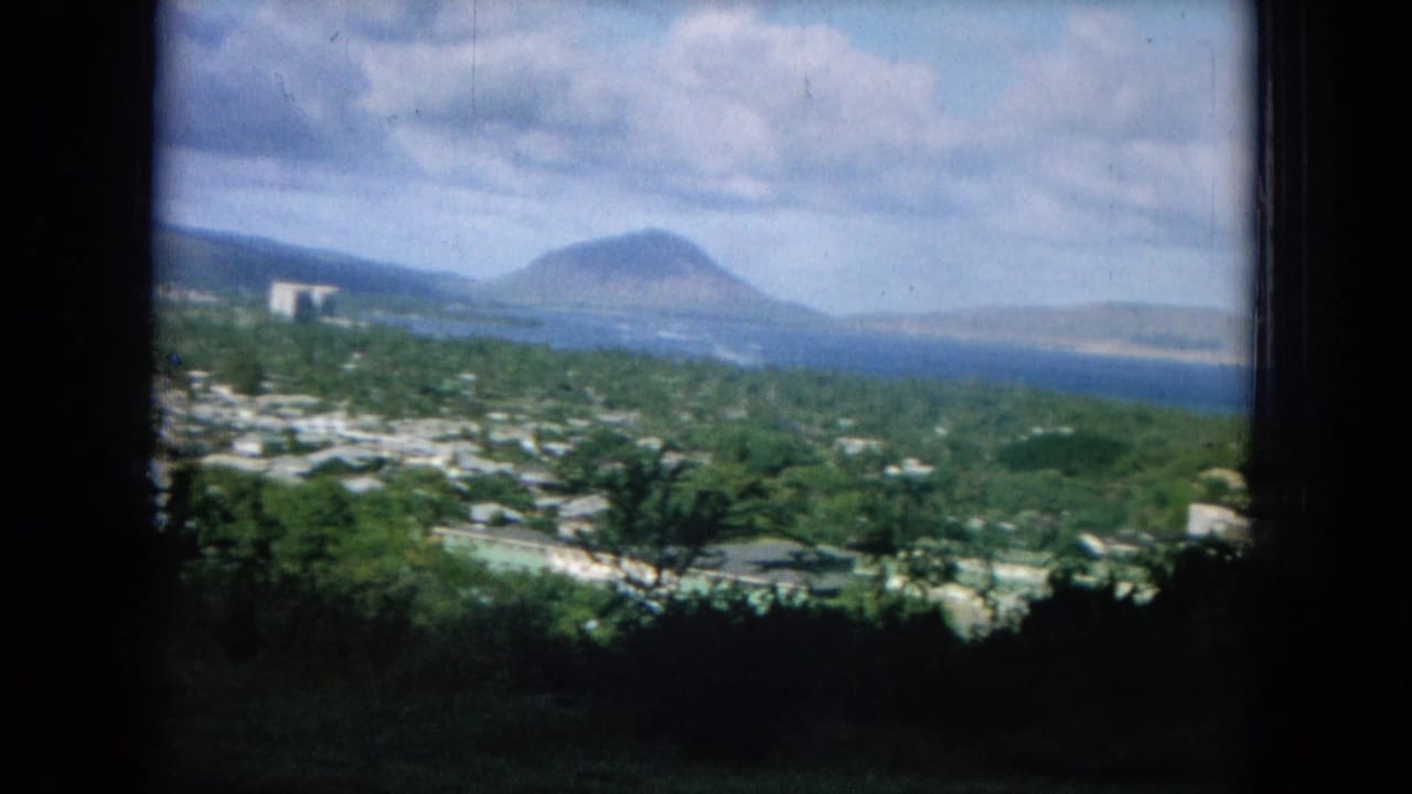Coastal panorama with trees, pale buildings, ocean, distant shoreline, and a rounded volcanic peak.