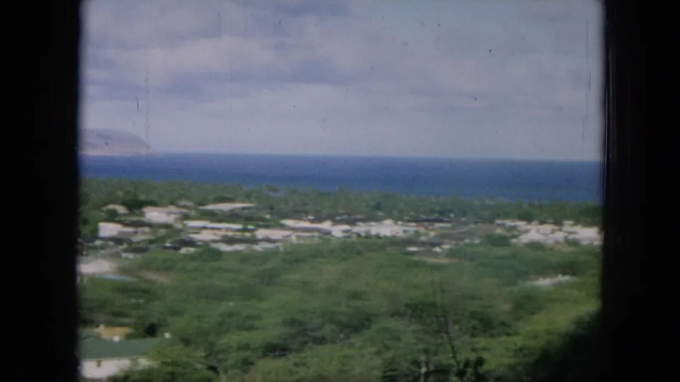 1964 8mm view from Oahu over green landscape, scattered low buildings, blue ocean, and a distant coastal landform.