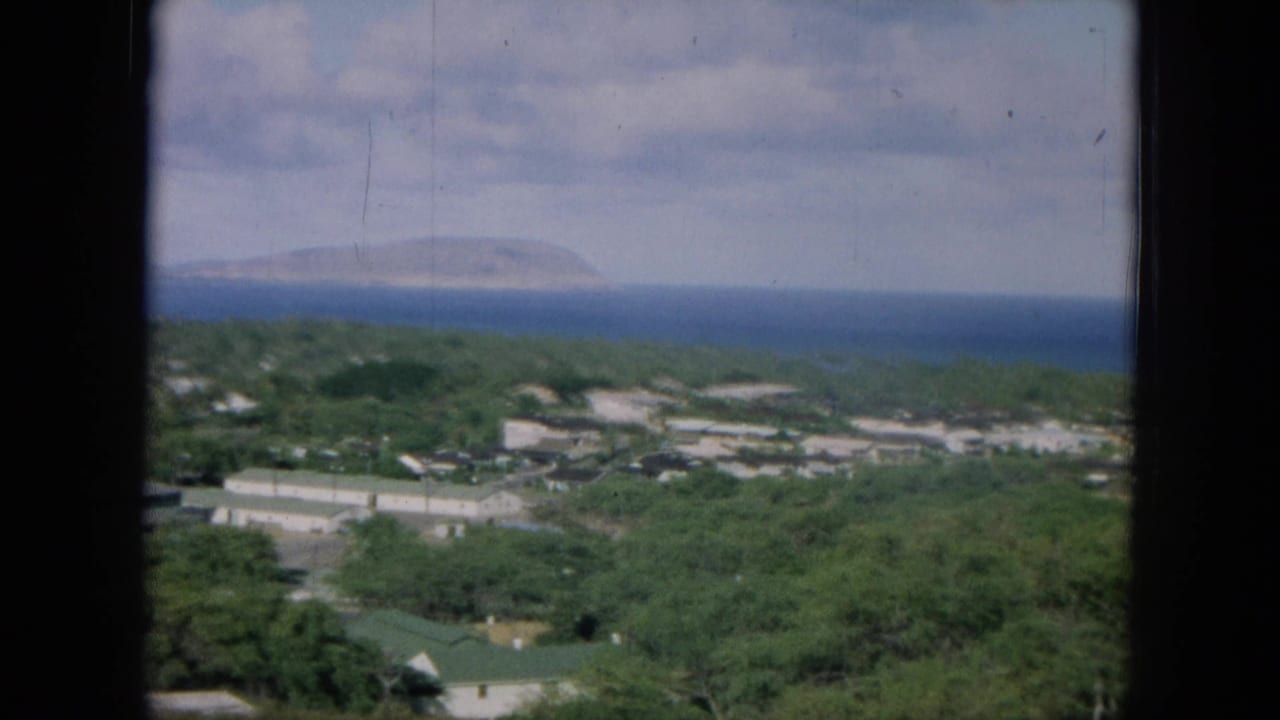 1964 Oahu home-movie frame showing low buildings amid trees, with deep blue ocean and a distant landform under clouds.