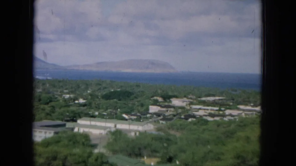 1964 8mm Oahu landscape frame with green tree cover, light industrial or residential buildings, blue water, and distant shoreline.