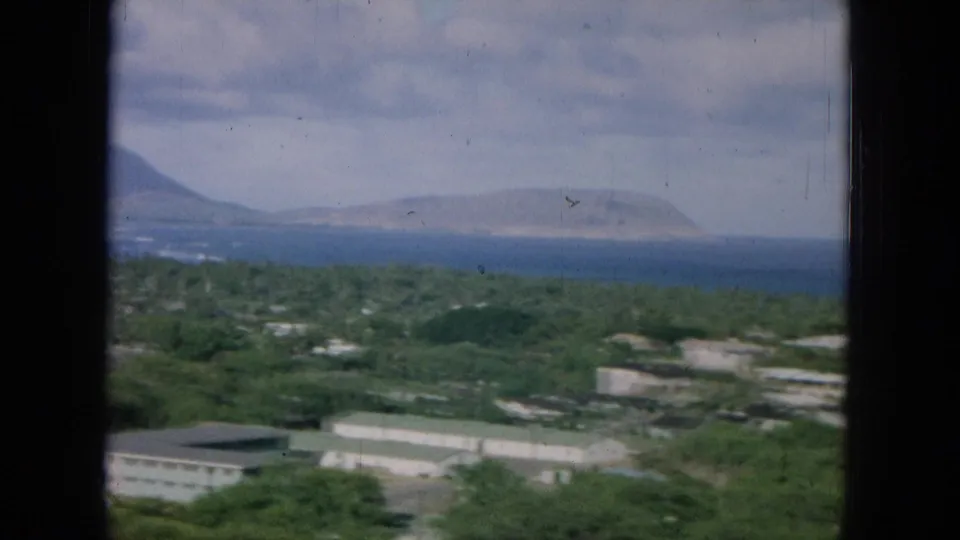 1964 Oahu 8mm frame showing a wide elevated view over trees and rooftops to blue ocean and a distant mountain-like landform.