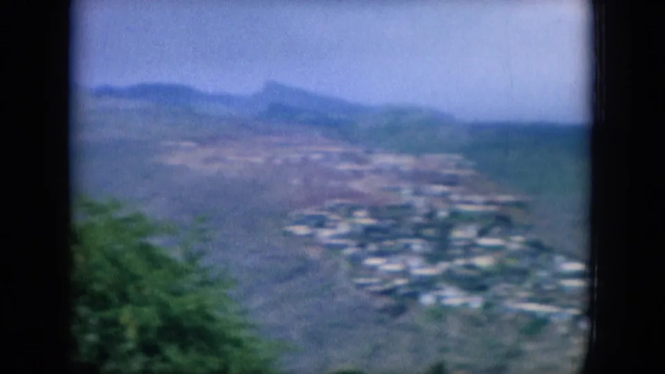 1964 Oahu home-movie frame showing clustered hillside houses, green vegetation, and distant mountain ridges under hazy light.
