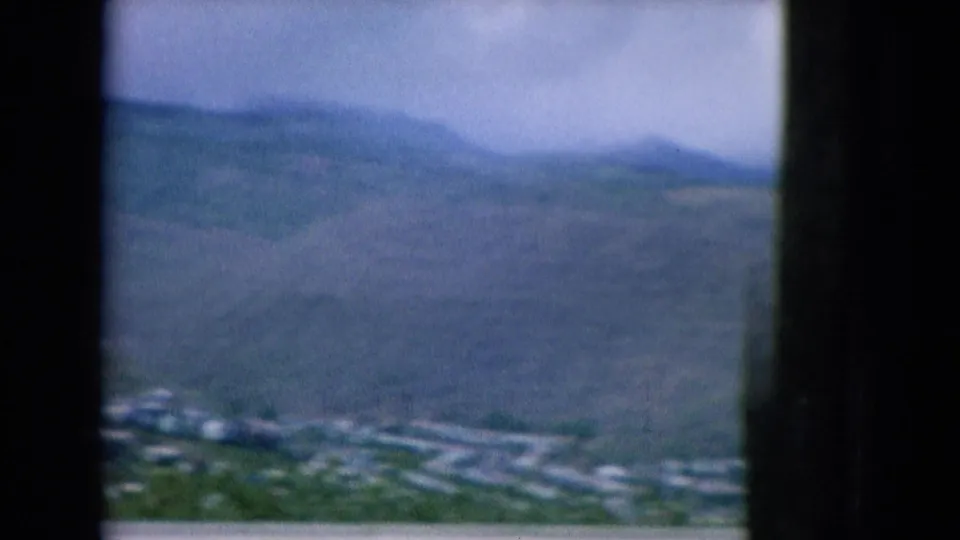 1964 Oahu frame showing a broad green hillside, scattered settlement, hazy sky, and dark side framing from the window view.