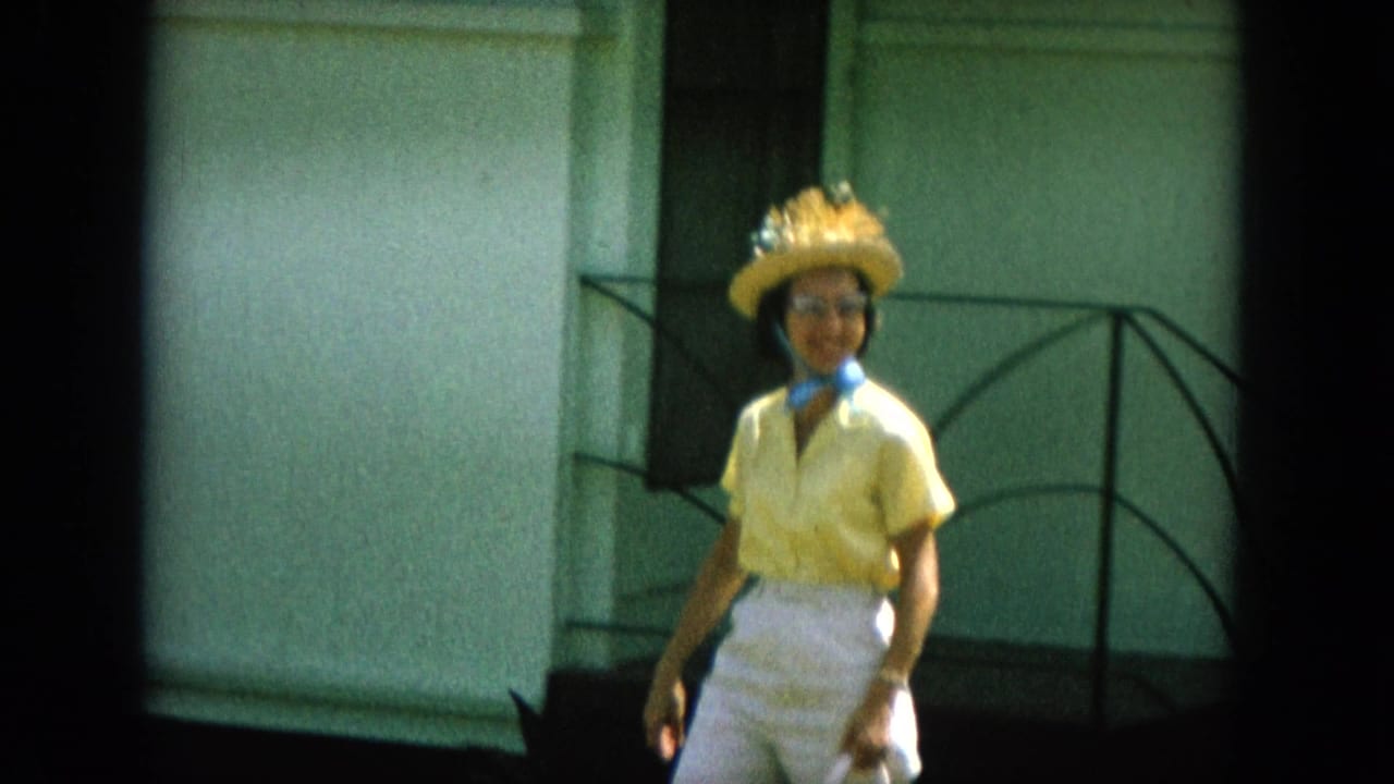 In 1964 Oahi, Hawaii footage, a woman in yellow clothing, straw hat, glasses, and scarf walks near a building exterior.