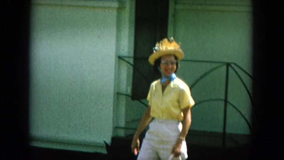 In 1964 Oahi, Hawaii footage, a woman in yellow clothing, straw hat, glasses, and scarf walks near a building exterior.