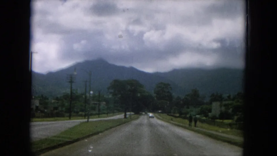 Passenger-view road footage in Oahi, Hawaii shows a paved road, roadside greenery, utility poles, and mountains beneath clouds.
