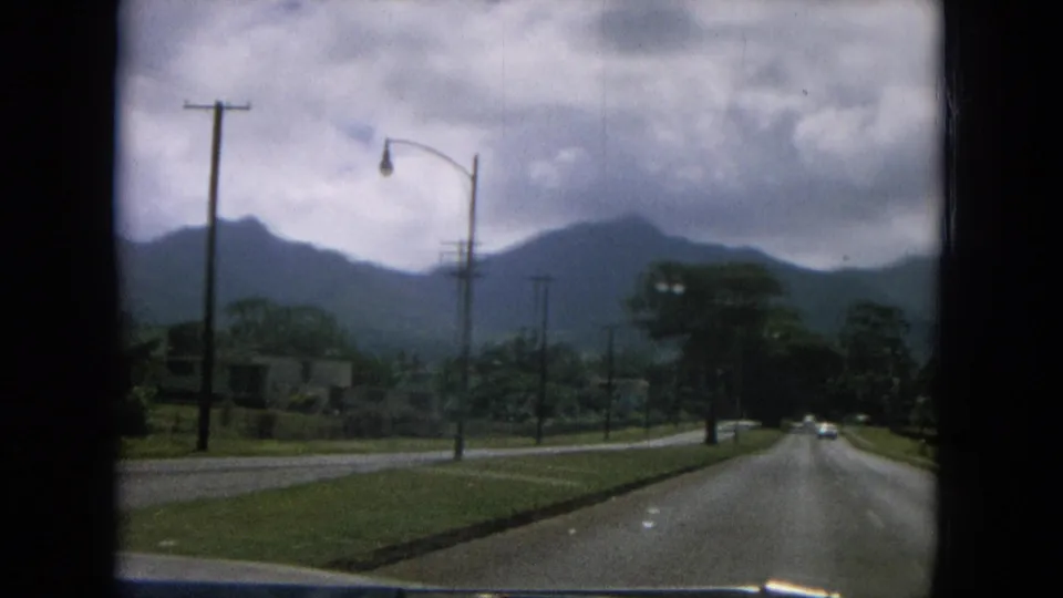 A 1964 home-movie road view shows a paved roadway, streetlight, utility poles, lush roadside, and dark mountains under cloud cover.