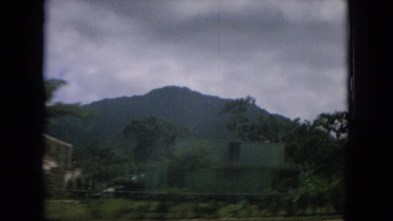 Mountain ridge behind trees and low roadside buildings under cloudy sky.
