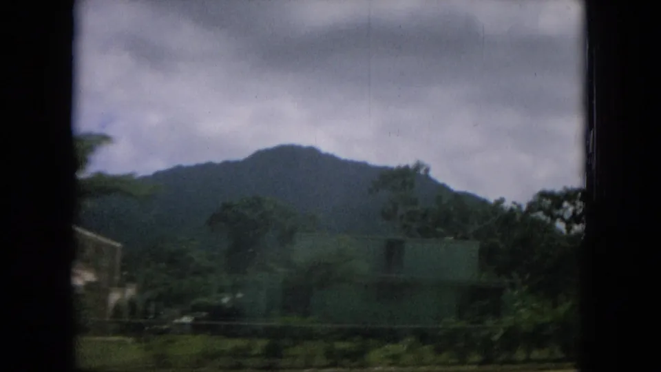 1964 Hawaii roadside frame with a low building, trees, green mountain ridge, and overcast sky seen from a moving vehicle.