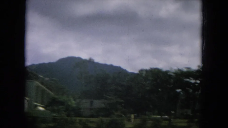 1964 Hawaii travel frame showing low roadside structures, trees, grass, and a mountain slope through soft 8mm motion blur.