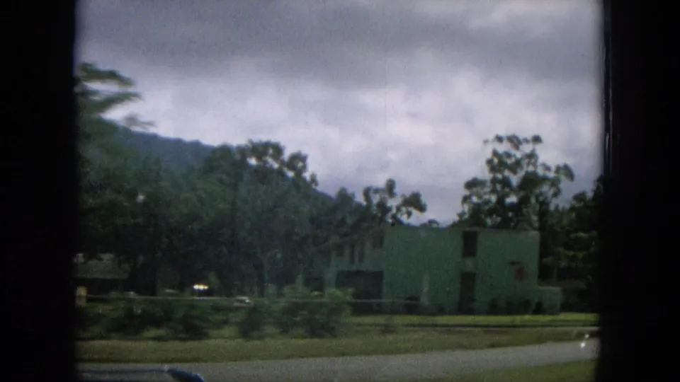 1964 Hawaii home-movie frame showing a pale roadside building, trees, grass, paved road, and cloudy mountain backdrop.