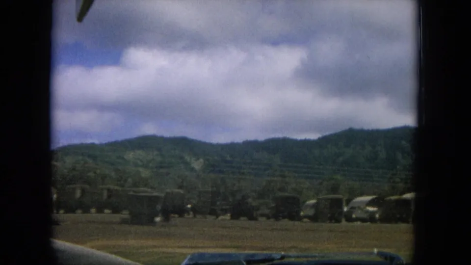 1964 8mm roadside view in Oahi, Hawaii, showing open land, low hills, cloudy sky, and passing foreground obstruction.