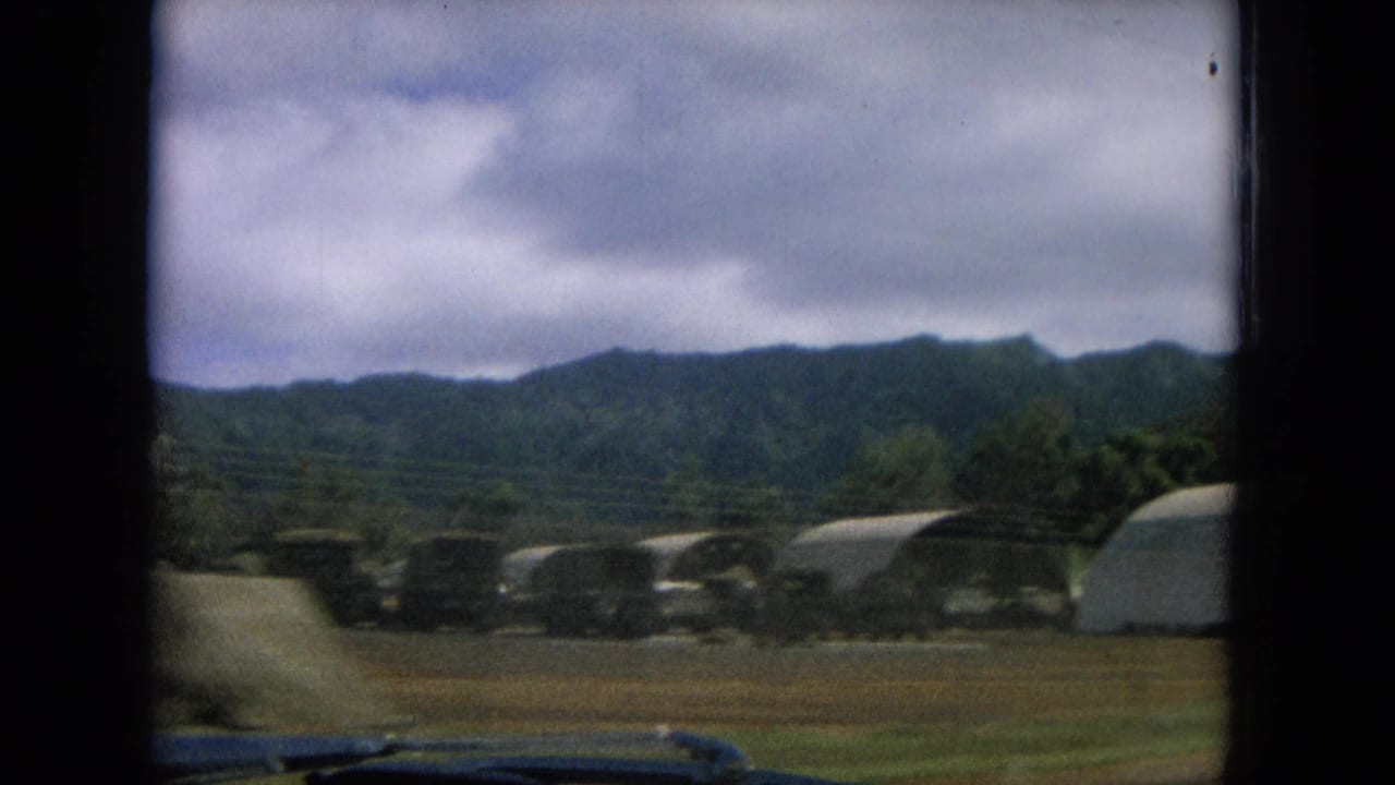1964 Oahi, Hawaii frame with arched hangar-like structures, open ground, trees, and green hills seen from a moving vehicle.