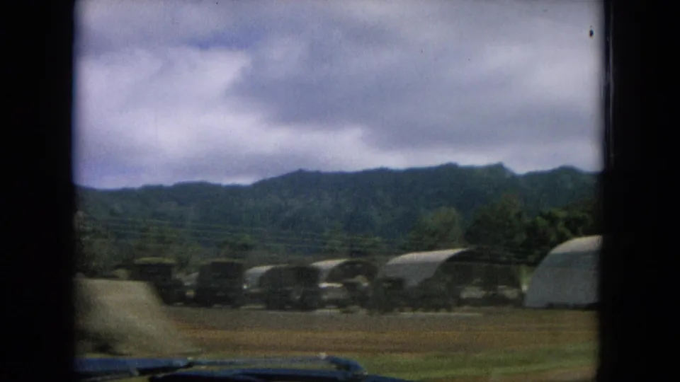 1964 Oahi, Hawaii frame with arched hangar-like structures, open ground, trees, and green hills seen from a moving vehicle.