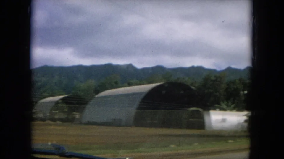 A 1964 8mm view in Oahi, Hawaii shows a large arched building near a smaller light-colored structure with hills behind.