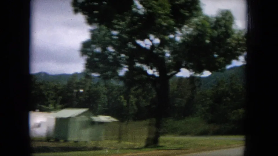 Final 1964 Oahi, Hawaii frame shows shed-like structures, a broad tree, grassy roadside, and distant hills from a moving car.