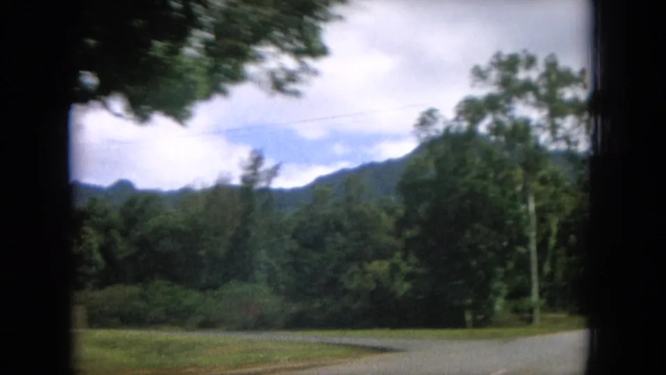 1964 8mm Hawaii home-movie view of a road beside trees and distant green mountains under a cloudy sky.