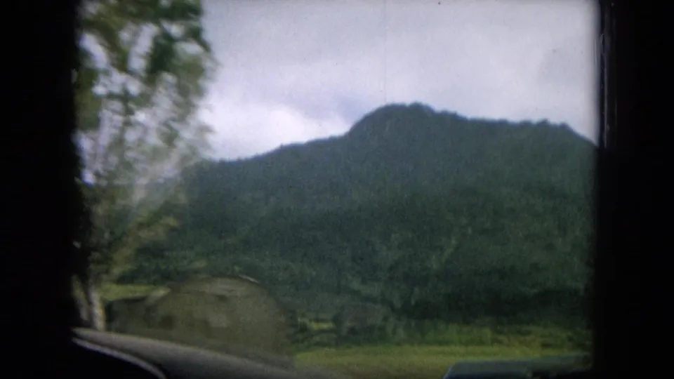 1964 Hawaii travel footage frame with green mountains, roadside plants, trees, and the lower edge of a car window or dashboard.