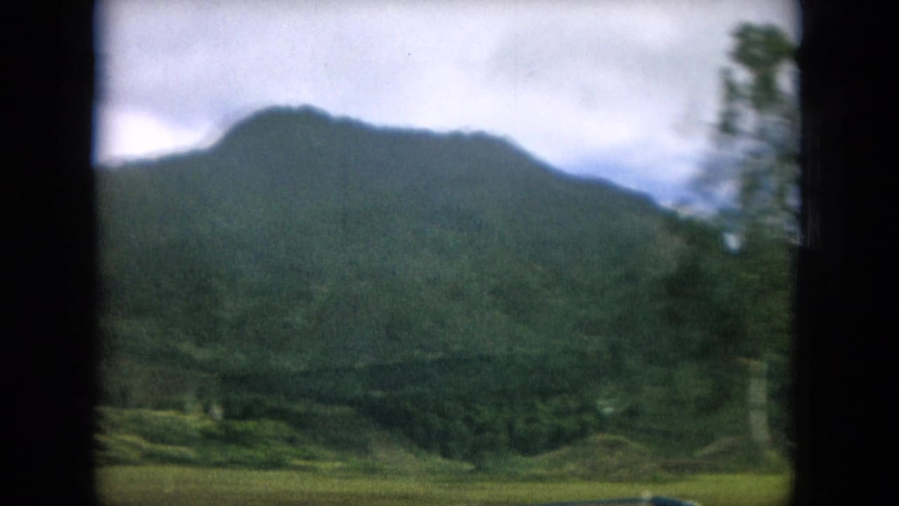 Rounded green mountain under cloudy sky with trees blurred at the roadside.