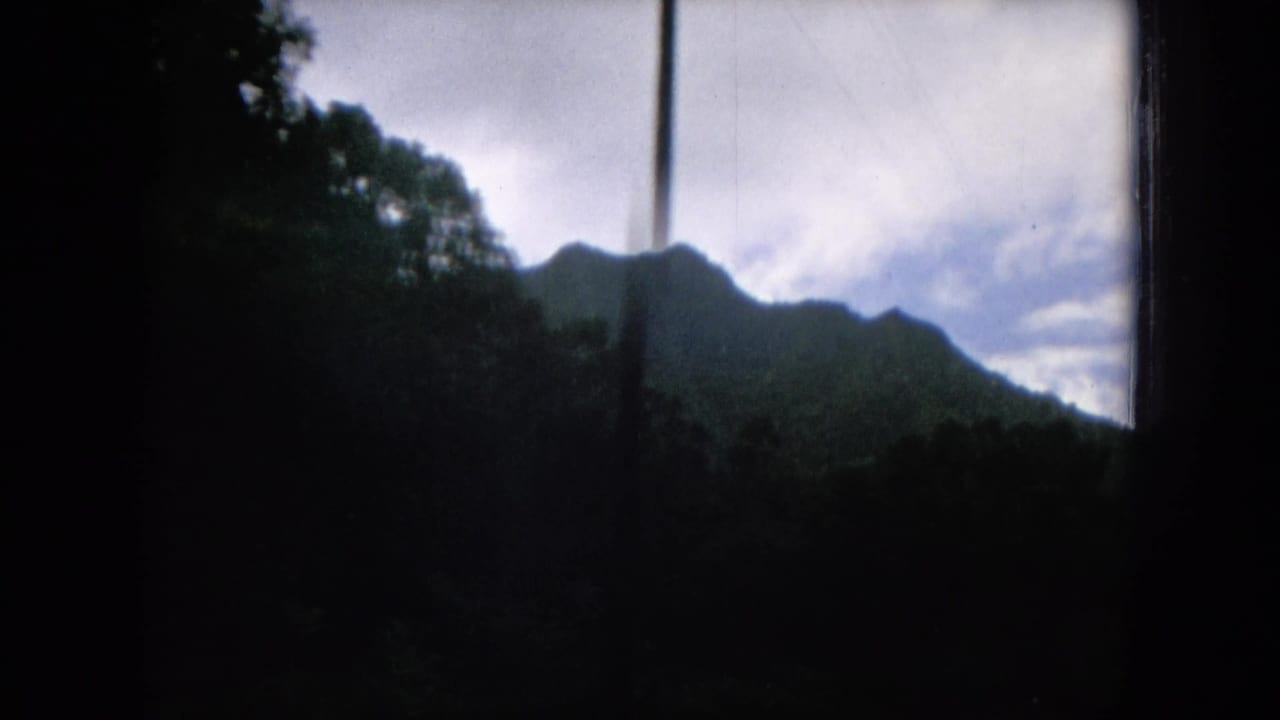 Dark vehicle window framing a green mountain ridge under a cloudy sky.