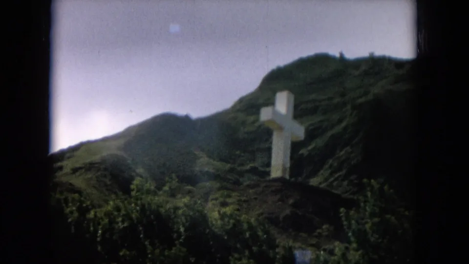 Distant 1964 Hawaii view of a white cross standing before steep, vegetation-covered mountain ridges.