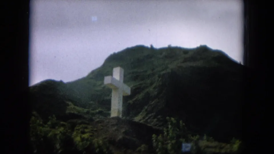 8mm home-movie frame of a white cross partly backed by a dark foreground slope and green Hawaiian mountain terrain.