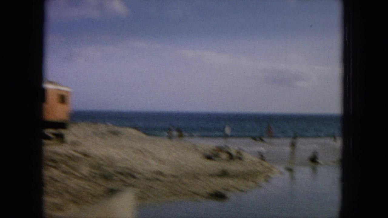 Coastal sand or roadside area seen through a dark vehicle window with water and small distant figures.