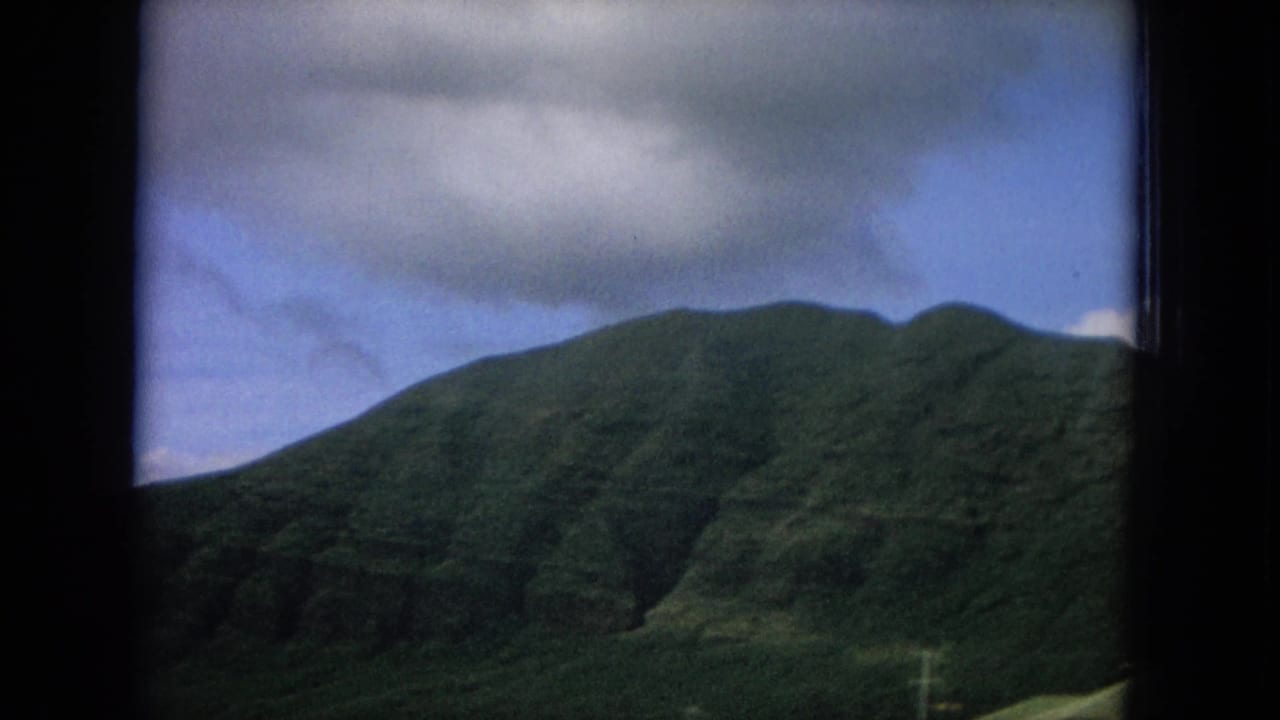Steep green mountain under a cloudy blue sky, framed by dark vehicle-window edges.