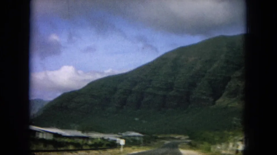 1964 8mm Hawaii roadside view with low homes or buildings at the foot of a large green mountain.