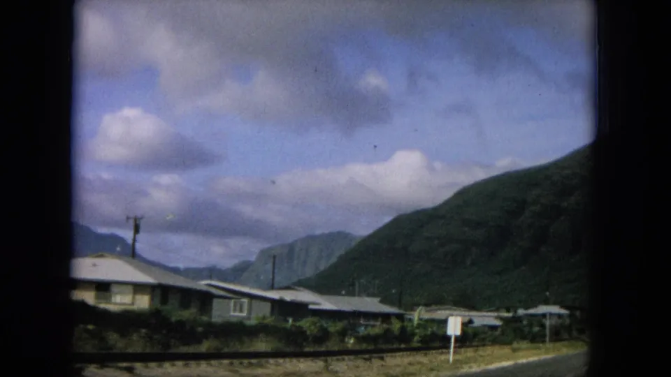 1964 8mm Hawaii home-movie frame showing small roadside homes, utility poles, and green mountains in the background.