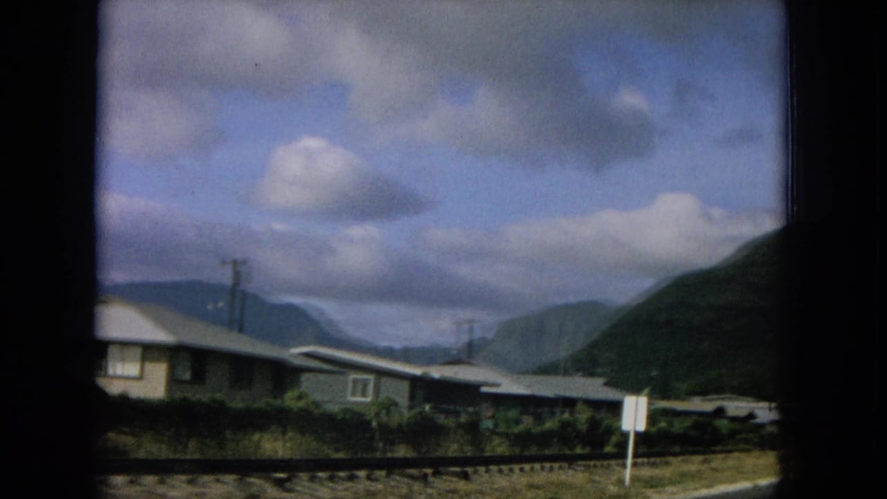 1964 Hawaii home-movie frame showing roadside houses, utility poles, mountains, and a small sign near a paved road.