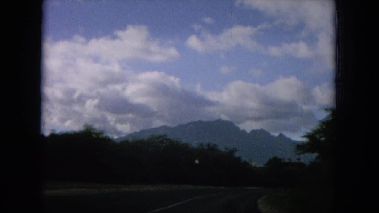 Moving-car view of a curving Hawaii road, roadside vegetation, distant mountain ridges, and a partly cloudy sky.