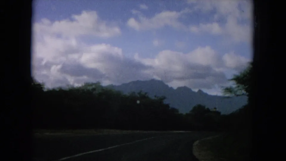 1960s 8mm view of a paved road running beside tropical trees, with mountain ridges and large clouds beyond.
