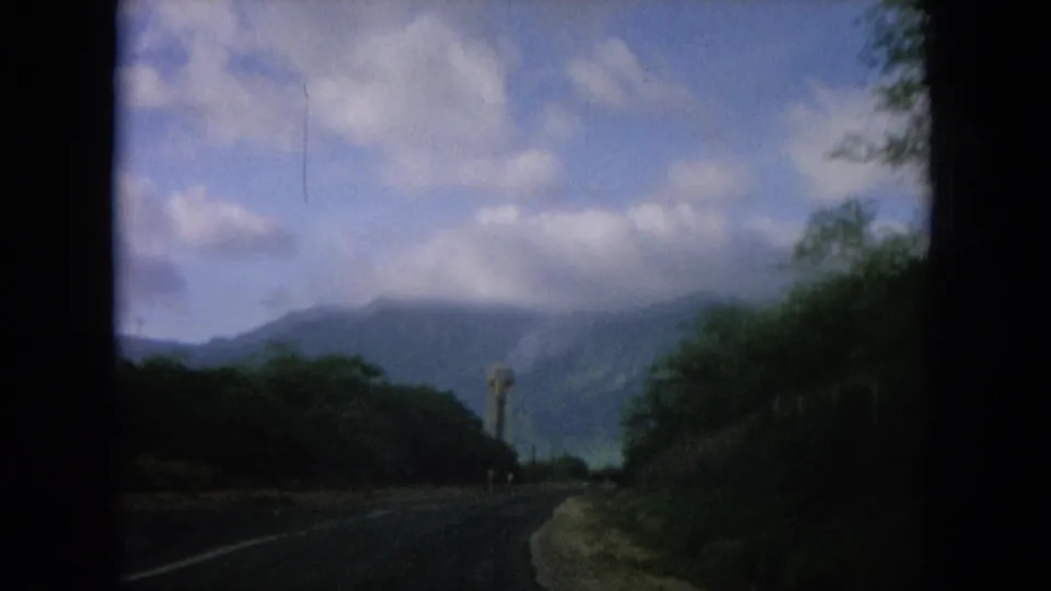 Hawaii road-travel frame with a utility pole, curving road edge, lush vegetation, and cloud-softened mountain slopes.